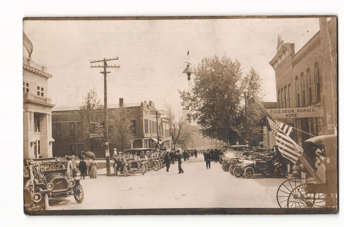 Farmington, Illinois Street View with Early Automobiles, Spectators, 1910 Postmark Postcard.: A sepia-toned photograph shows a street scene with early 20th-century automobiles lining both sides. People in period attire stand on sidewalks and in the street. Two to three-story brick and stone bu