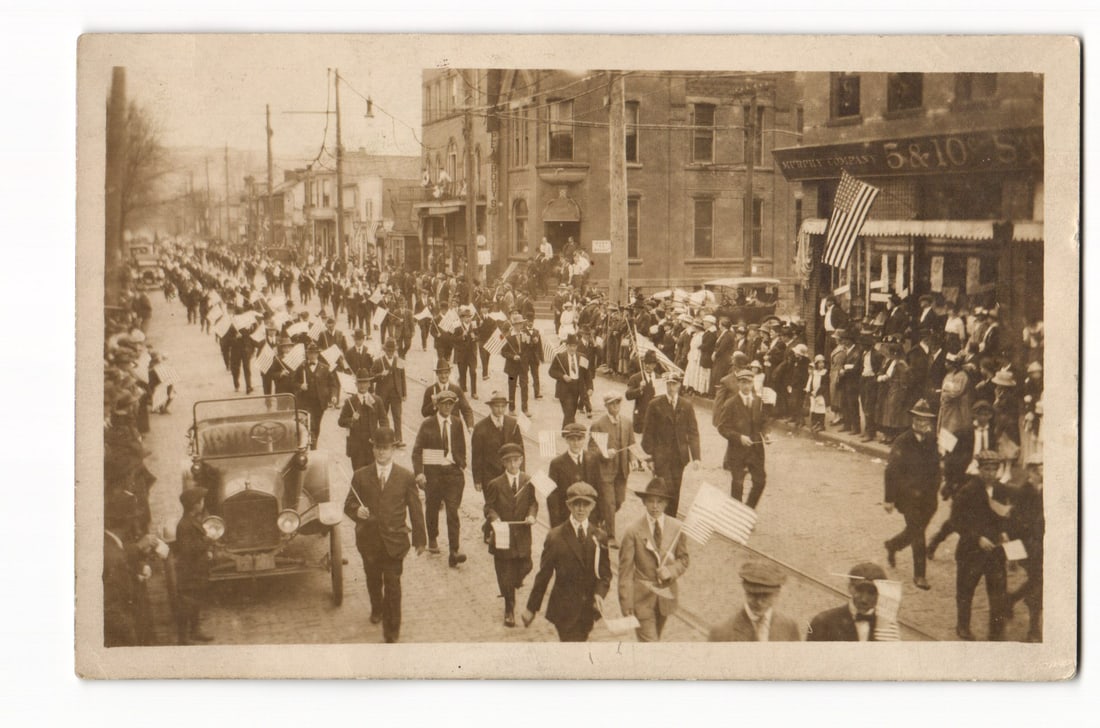 Newport, N.H. Real Photo Postcard of Early 20th Century Street Parade with American Flags: A sepia-toned Real Photo Postcard (RPPC) depicts a large procession moving along a cobblestone street. Participants, predominantly men in suits and hats, are visible, with many carrying American flags