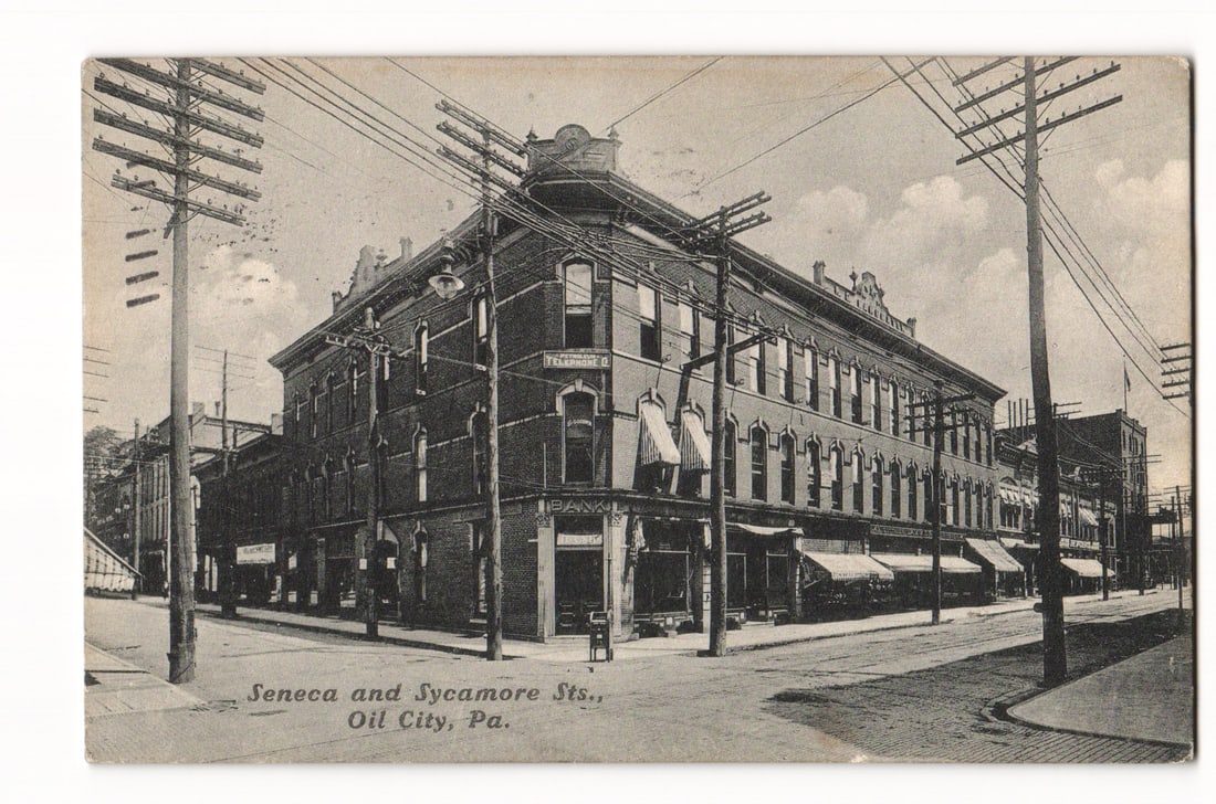 Oil City, PA Postcard: Seneca & Sycamore Sts. Corner View, Bank, Utility Poles, circa 1909: A sepia-toned photographic print depicts a street corner scene. The primary subject is a multi-story brick commercial building at an intersection, viewed from a low angle. The building features arched