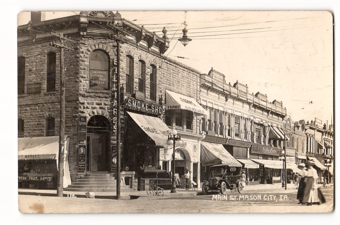 Main St. Mason City, IA, Street Scene with Businesses & Early Automobile, RPPC Postcard c. 1914: A sepia-toned photograph shows Main Street, Mason City, Iowa, from a corner. The prominent two-story left corner building of rough-cut stone has arched second-floor windows. Signage includes "LAW OFFI