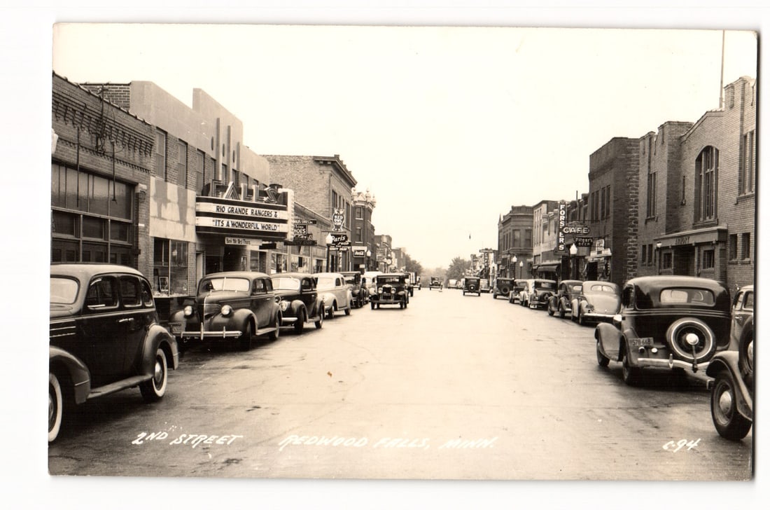 Redwood Falls, Minn. 2nd Street with Vintage Cars & "It's a Wonderful World" Marquee Postcard: A black and white photograph depicts a street view identified by printed text at the bottom: "2ND STREET REDWOOD FALLS, MINN.". The perspective looks down the street, with numerous automobiles from th