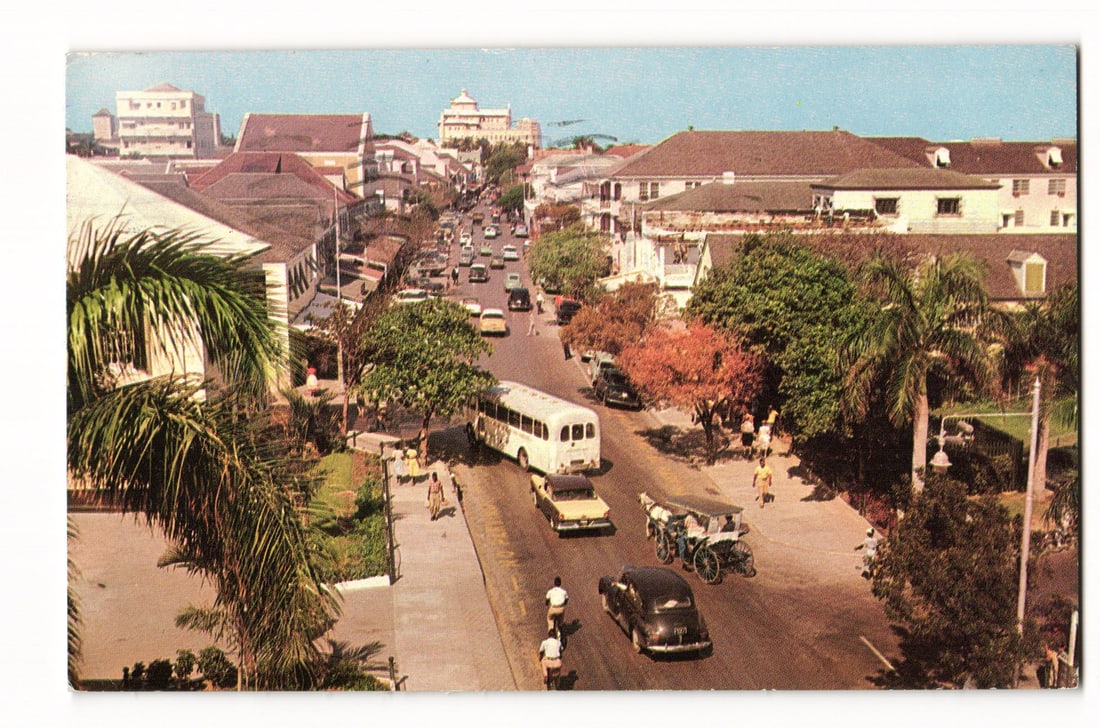 Nassau, Bahamas: Elevated View of Bay Street with Traffic & Buildings, c. 1953 Colour Postcard: Elevated color photo: Bay Street, Nassau (per reverse). Multi-story buildings, awnings. Mid-20th C. cars, bus, carriage. Pedestrians. Tropical flora, palms. Hazy sky. Divided back. Features one circul
