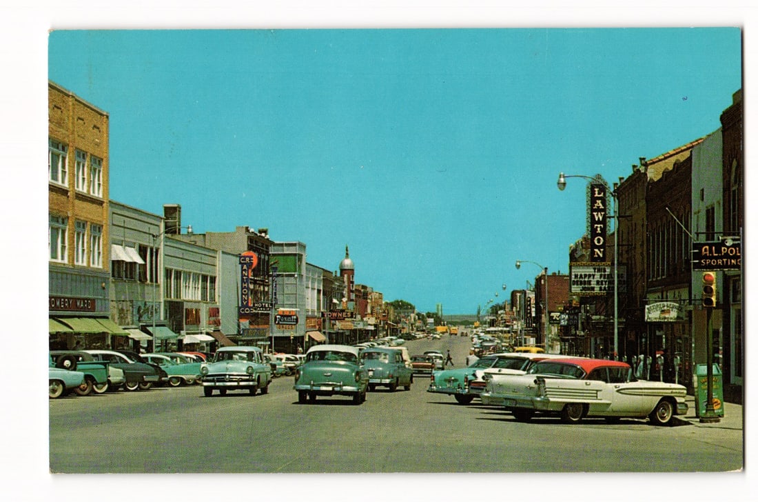 Street Scene in Lawton, Oklahoma, with 1950s Cars and Storefronts, Colorchrome Postcard: A color photograph depicts a street scene in Lawton, Oklahoma, looking down a wide thoroughfare lined with parked automobiles, characteristic of the 1950s. Buildings on the left feature signage for "M