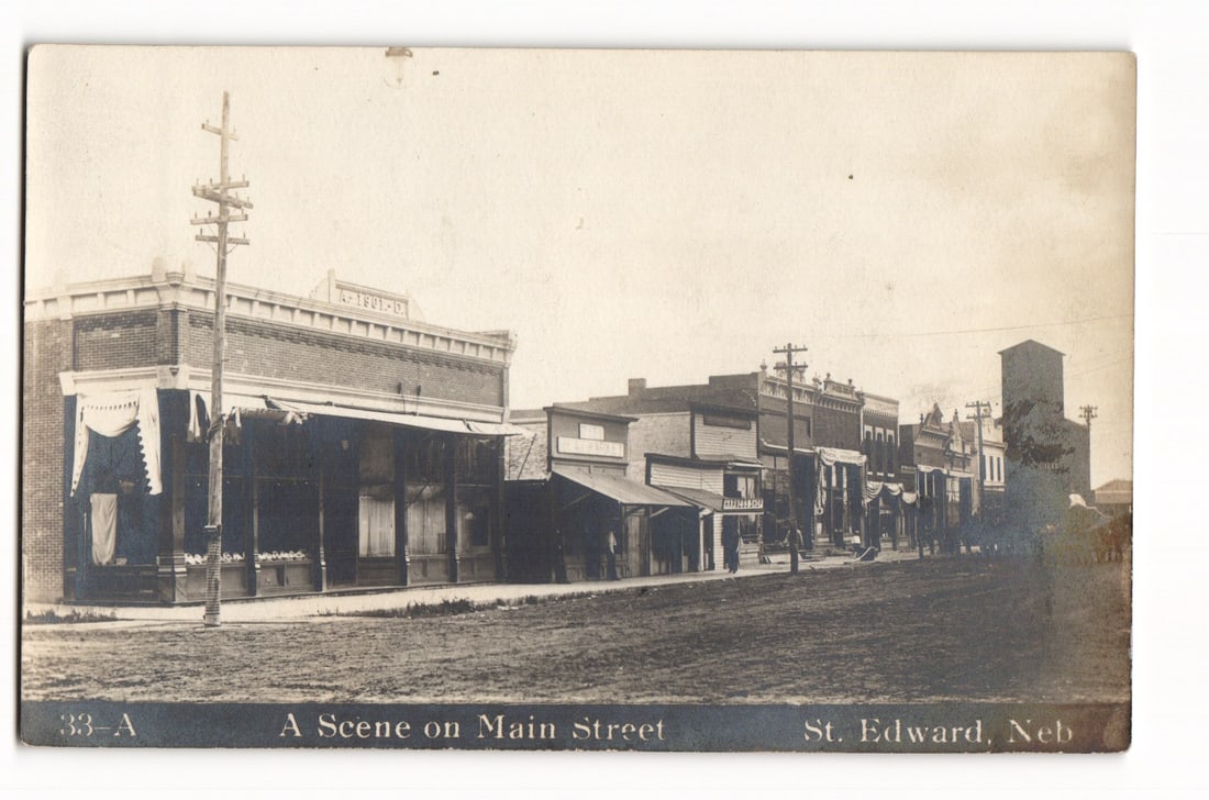 Real Photo Postcard: Main Street view, St. Edward, Neb. featuring A.F. & A.M. 1907 building.: A sepia-toned photograph shows an angled perspective of commercial buildings along an unpaved Main Street in St. Edward, Nebraska. This is a Real Photo Postcard (RPPC). Utility poles line the street.