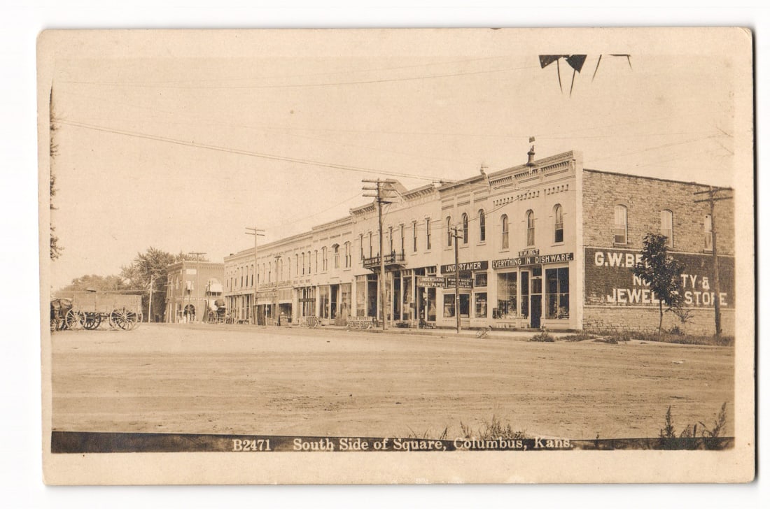 Columbus, Kansas, South Side of Square Street View with Businesses, Early 20th C. RPPC postcard: A sepia-toned Real Photo Postcard presents a view of the South Side of the Square in Columbus, Kansas. The image depicts a row of predominantly two-story commercial buildings with various architectura