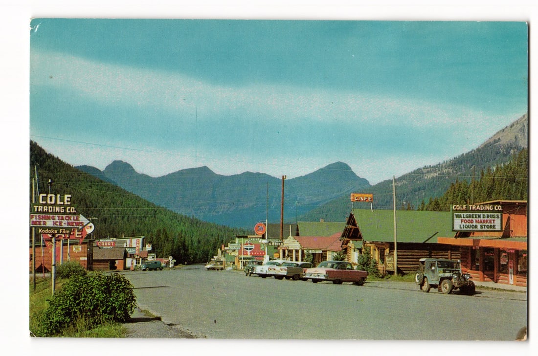 Cooke City, Montana: Cole Trading Co., Northern Yellowstone Entrance, vintage street view postcard: The color photograph depicts a street scene in Cooke City, Montana, looking down a paved road. On either side of the road are one and two-story wooden commercial buildings and several parked vintage a