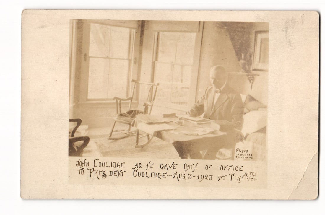 John Coolidge Giving Oath of Office to President Coolidge, Plymouth VT, 1923 RPPC Postcard: A sepia-toned Real Photo Postcard (RPPC). The image depicts a man seated at a table covered with papers in an interior setting. He is dressed in a suit and tie and appears to be engaged with the docum