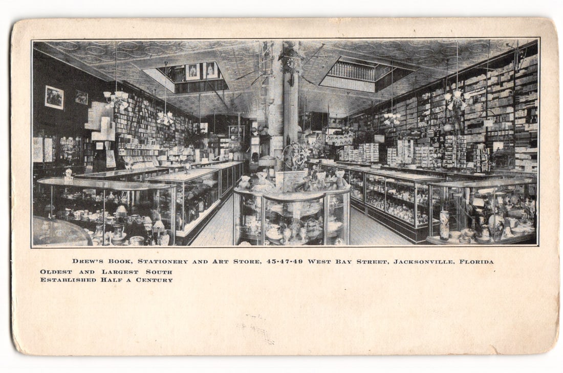 Drew's Book, Stationery & Art Store, Jacksonville, FL, 'Established Half A Century' postcard: A black and white printed photographic interior view of Drew's Book, Stationery and Art Store. The perspective is looking down a central aisle, flanked by glass display cases on the left, right, and i