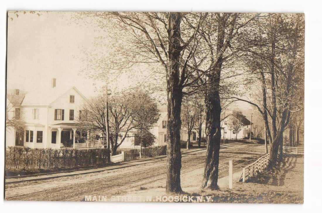 Main Street, N. Hoosick, N.Y. Early 20th C. Residential Street Scene, Real Photo Postcard: A sepia-toned photograph depicts a view looking down an unpaved street, identified as Main Street in N. Hoosick, N.Y. Several multi-story residential buildings line the left side, including a prominen