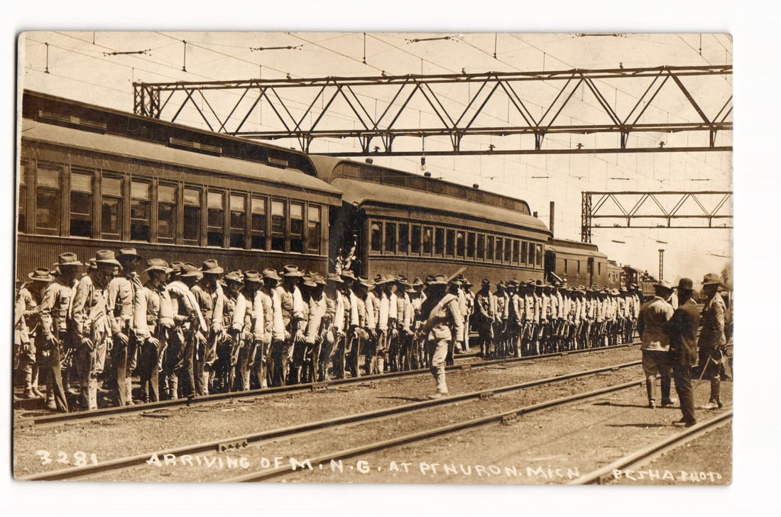 Arrival of Michigan National Guard (M.N.G.) at Pt. Huron, Mich. by Train, Sepia RPPC Postcard: A sepia photograph depicts numerous soldiers in uniform and hats in formation by a multi-car passenger train. In the foreground are railway tracks, and in the background is an overhead metal gantry st