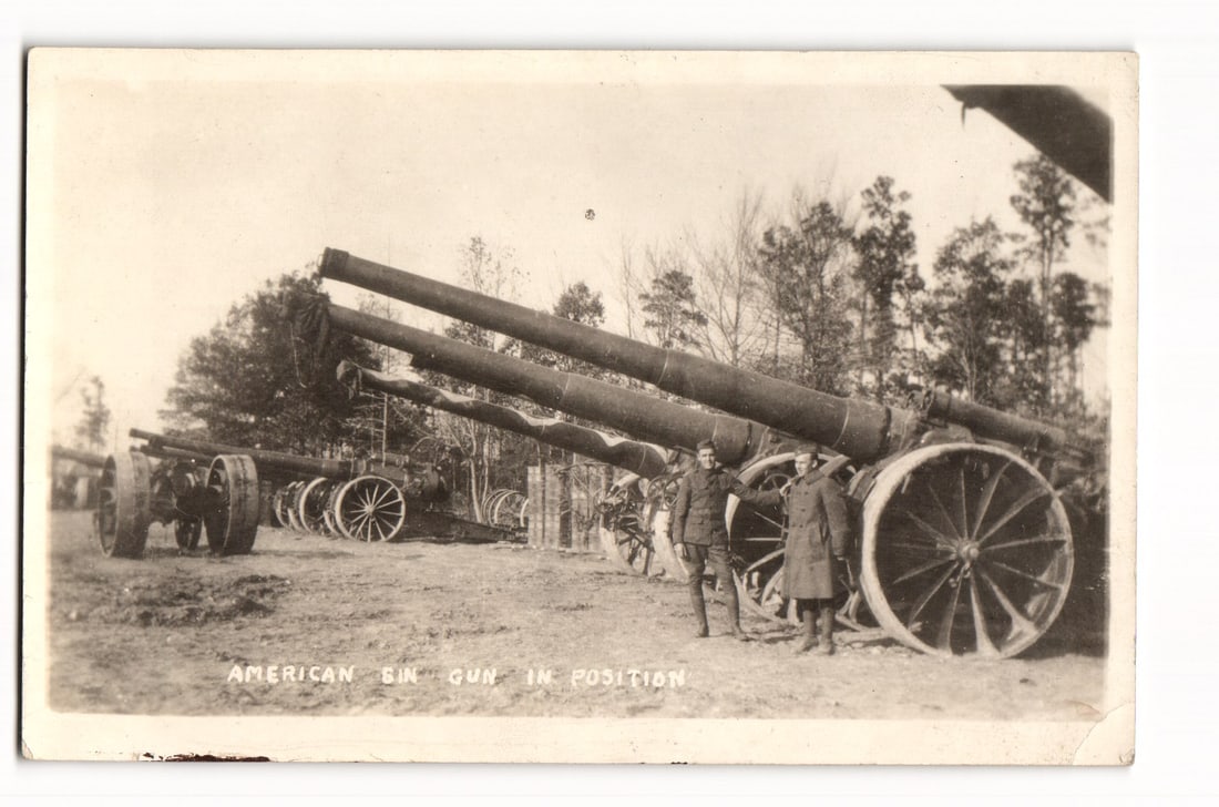 American 8-inch Guns in Position with Soldiers, Early 20th Century Military Real Photo Postcard: A sepia-toned photograph depicts a row of large artillery pieces, identified by a printed caption as "AMERICAN 8IN GUN IN POSITION". Two soldiers in uniform, wearing long coats, leggings, and service
