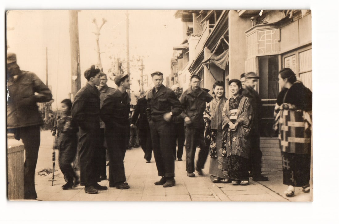 Real Photo Postcard of Soldiers and Local Women in Kimonos on a Street, Mid-20th Century Japan: A sepia-toned photographic image depicts a street scene. In the foreground and middle ground, a group of at least seven men, several in military-style uniforms including garrison caps and service atti
