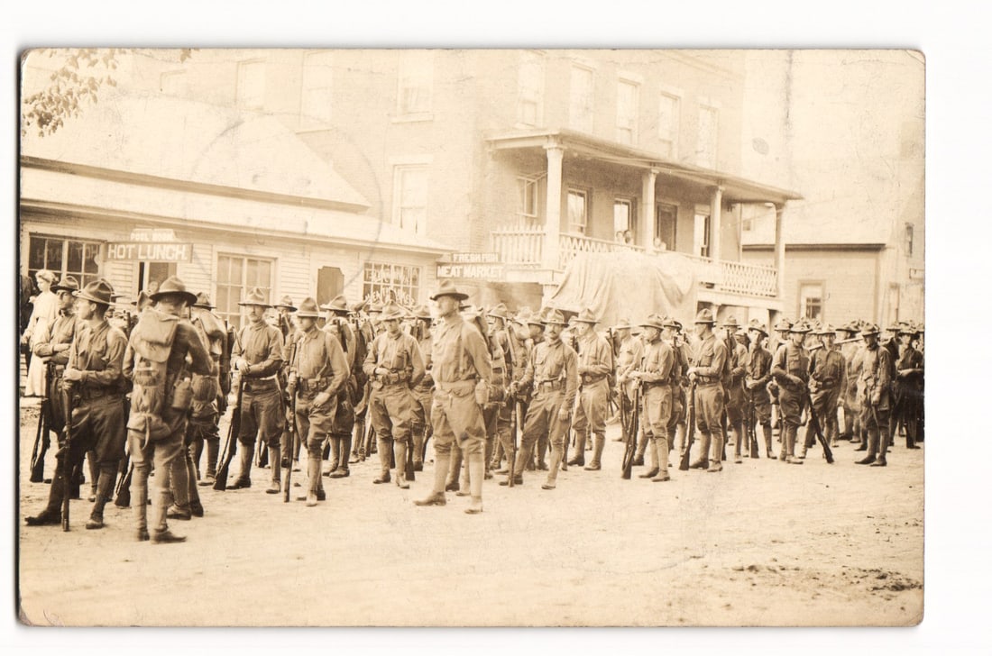 Real Photo Postcard: Soldiers in Formation, Rouses Point, N.Y., The Grand Union Meat Market, 1915.: A sepia-toned photograph depicts a large group of soldiers in early 20th-century military uniforms, standing in formation on an unpaved street. Many soldiers are equipped with rifles and backpacks, an