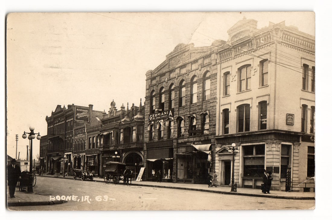 Boone, Iowa Street Scene with Germania & Hans Bros. Buildings, Early 20th C. RPPC Postcard: A sepia-toned photographic image captures an angled street view in Boone, Iowa. On the right side of the unpaved street, a row of multi-story commercial buildings recedes into the distance. Prominent