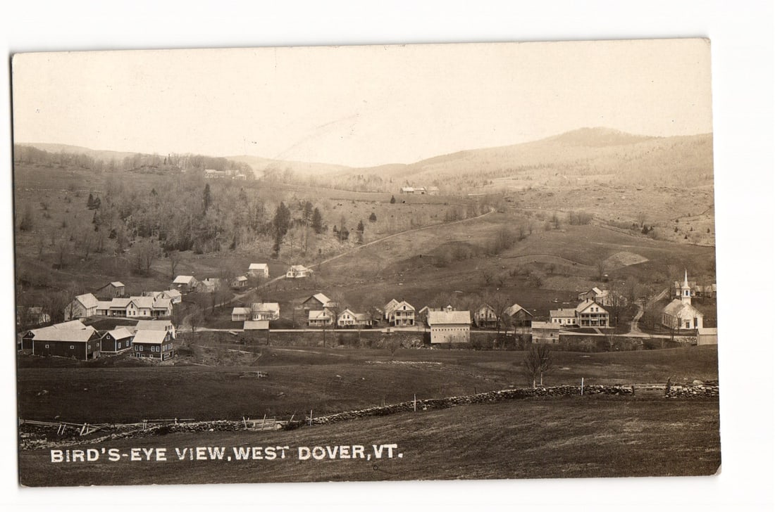 Bird's-Eye View of West Dover, VT, Depicting Village and Hills, RPPC Postcard, circa 1910s: A sepia-toned photographic image captures a bird's-eye view of West Dover, Vermont. The village, comprised of various wooden buildings including houses, barns, and a church with a steeple on the far r