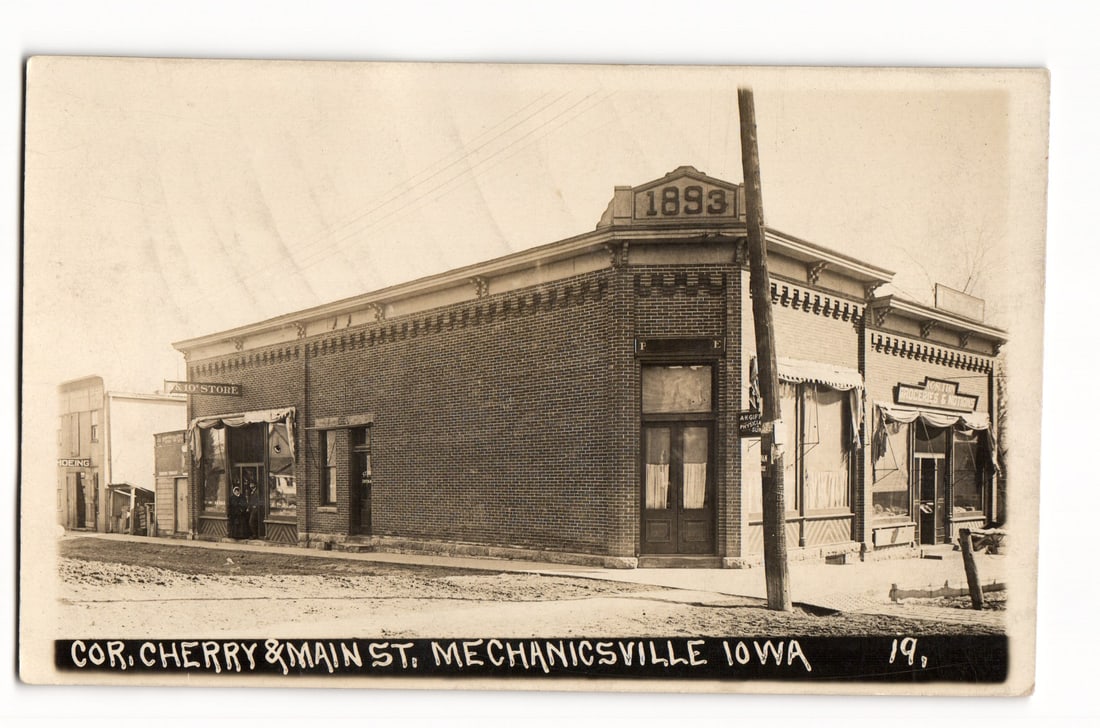 RPPC Postcard: Corner of Cherry & Main St., Mechanicsville, Iowa, "1893" Building Scene: A sepia-toned photographic image captures a street corner scene. The primary subject is a brick commercial building, viewed from an angle, with "1893" prominently displayed on its cornice. The buildin