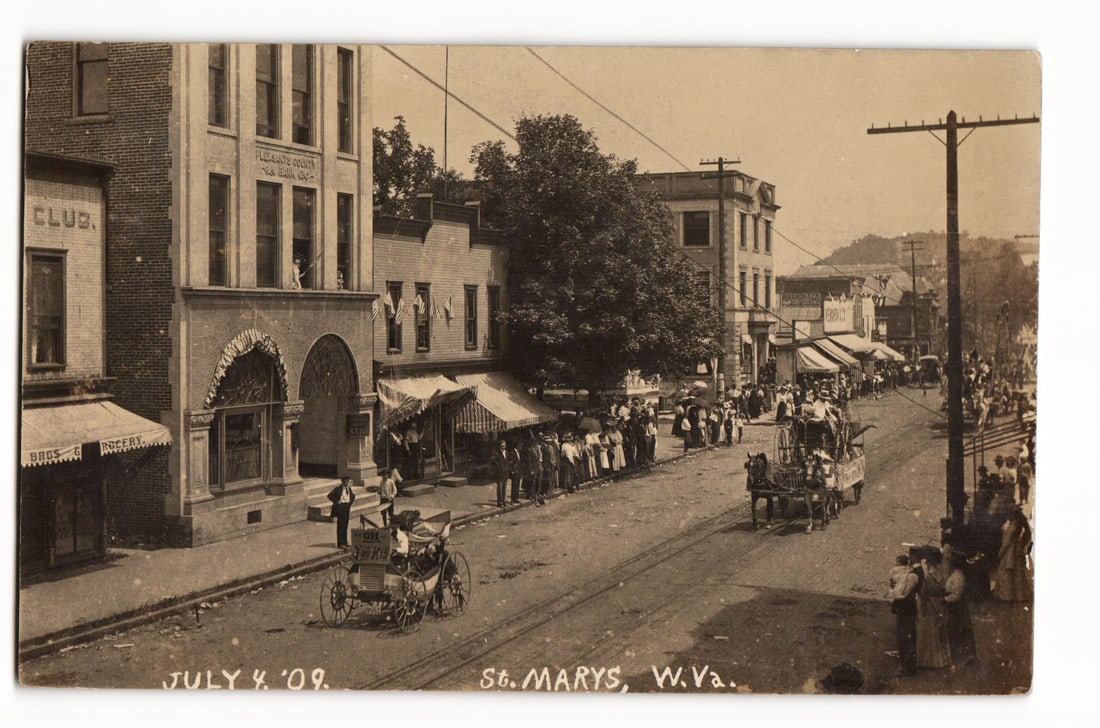 July 4, 1909 Parade, St. Marys, W.Va., "The Bird Sellers," Street View, RPPC Postcard: Sepia-toned photograph of a street scene in St. Marys, W.Va., dated July 4, 1909. Crowds line the unpaved street, featuring trolley tracks. A horse-drawn vehicle in the foreground displays a sign "THE