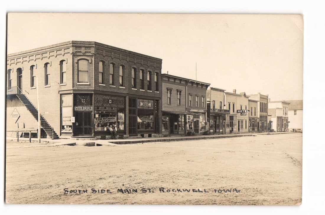 South Side Main St., Rockwell, Iowa, Businesses including Smith Drugs, Hotel Coye, RPPC Postcard: South Side Main Street in Rockwell, Iowa, is depicted in a photographic image. This Real Photo Postcard (RPPC) shows a row of commercial buildings lining an unpaved street. On the left, a prominent tw