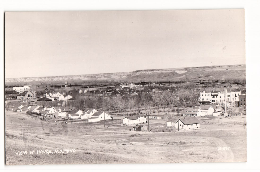 Panoramic View of Havre, Montana, Showcasing Residential and Commercial Buildings, RPPC Postcard: The image is a sepia-toned panoramic photograph taken from an elevated perspective, looking down over the town of Havre, Montana. In the foreground, a sparsely vegetated hillside slopes downwards towa