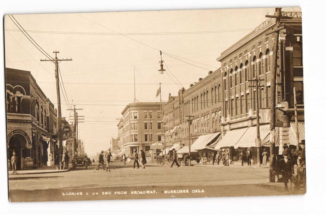 Muskogee, Oklahoma: Looking South on 2nd from Broadway. Early 20th C. Real Photo Postcard.: A sepia-toned photograph captures a street view in Muskogee, Oklahoma, looking south on 2nd Street from Broadway. Multi-story brick commercial buildings with awnings line the street. Signage on the ri