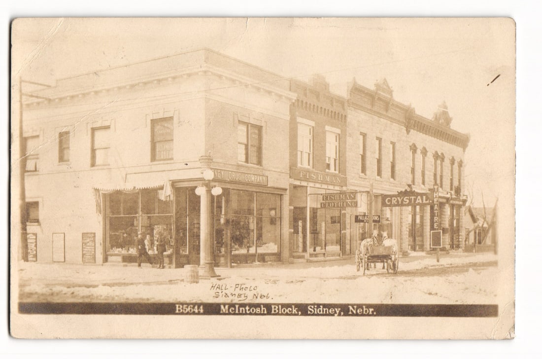 RPPC McIntosh Block, Sidney, Nebr. showing storefronts, Crystal Theatre, 1913 Postcard: A sepia-toned photographic street scene depicts the McIntosh Block in Sidney, Nebraska. The image shows an angled perspective of two-story brick buildings lining a snow-covered street and sidewalks. P