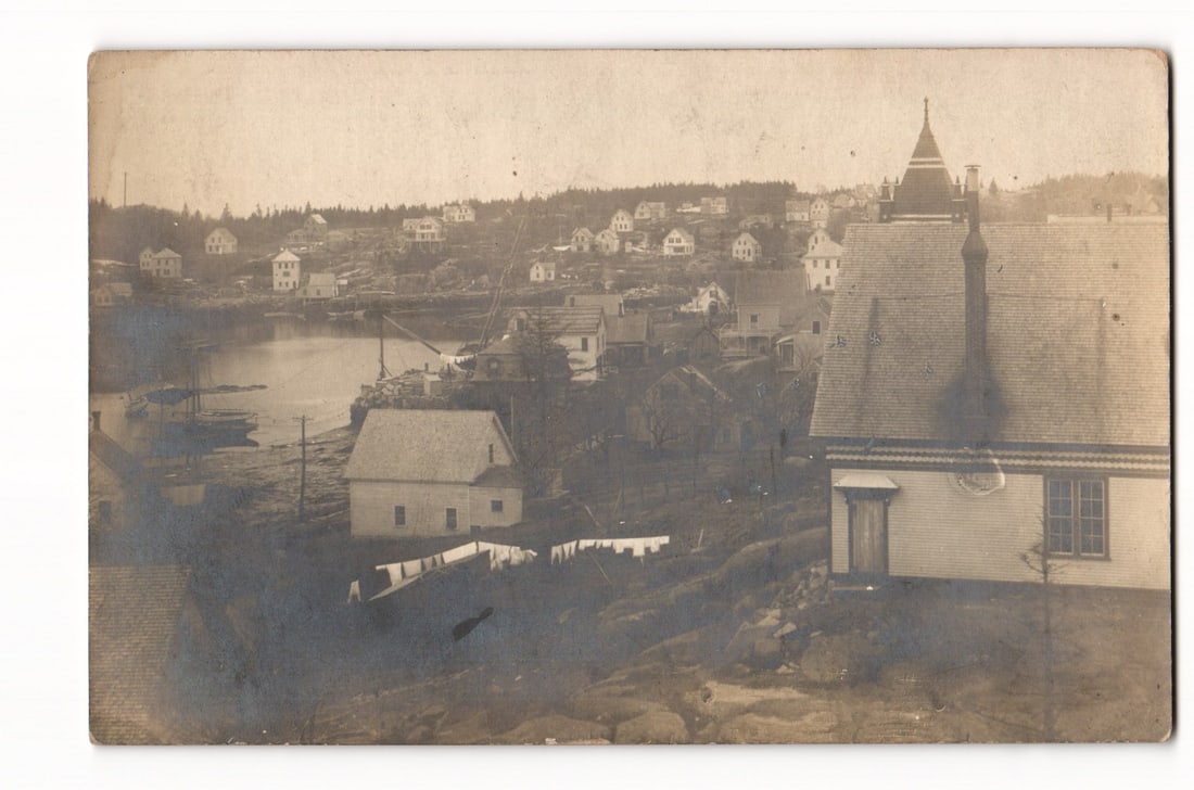 RPPC Sepia Photograph Coastal Village Houses Boats Harbor Scene Undivided Back Postcard c. 1900s: A sepia-toned photographic view depicts a coastal village with numerous wooden houses clustered on a hillside sloping down to a body of water, likely a harbor or bay. Several small boats are visible m