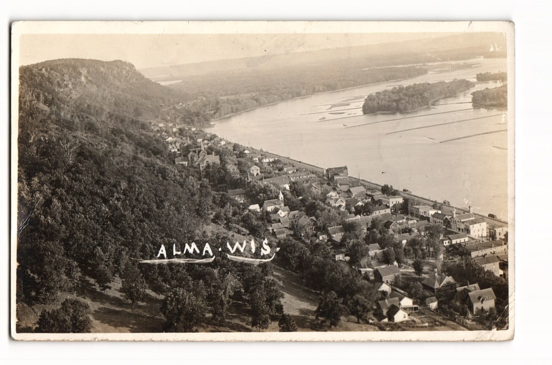 Alma, Wisconsin, Elevated View of Town and Mississippi River, 1920 Real Photo Postcard: A high-angle photographic view shows the town of Alma, Wisconsin, nestled between a steep, tree-covered bluff on the left and a wide river, identified as the Mississippi on the reverse, on the right. 