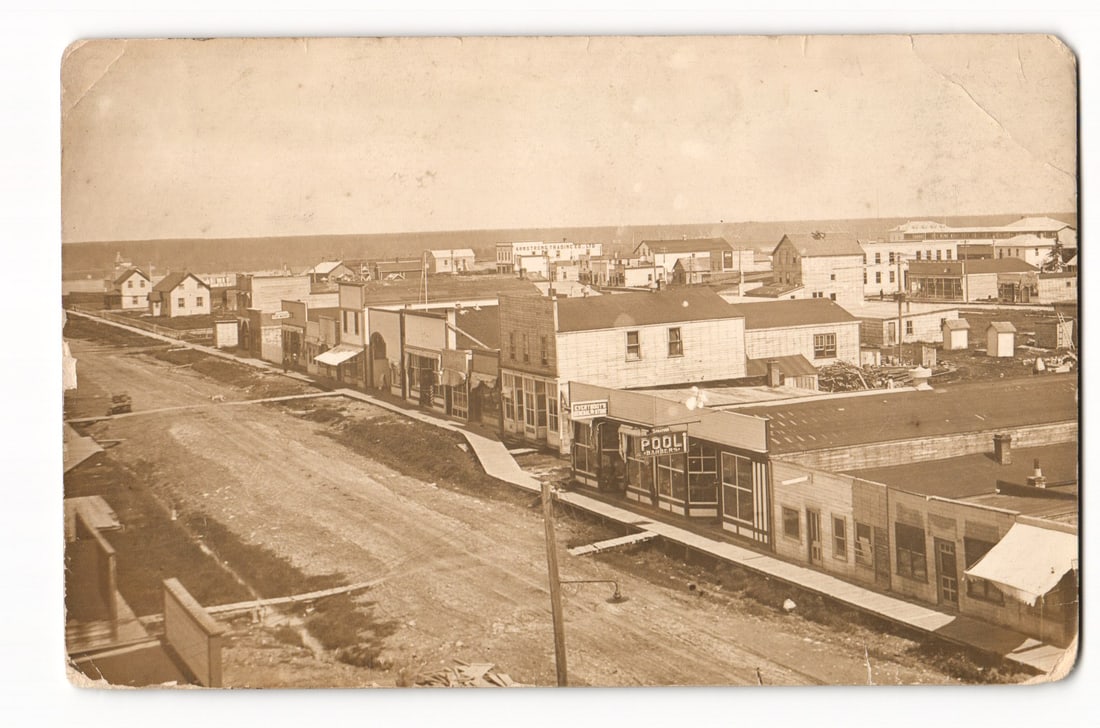 RPPC Postcard: High-Angle View of Early 20th Century Canadian Town with Pool Room & Restaurant: A sepia-toned photographic image presents a high-angle, panoramic view of a town. The perspective looks down onto unpaved dirt roads and wooden boardwalks lining commercial streets. Numerous wooden bu