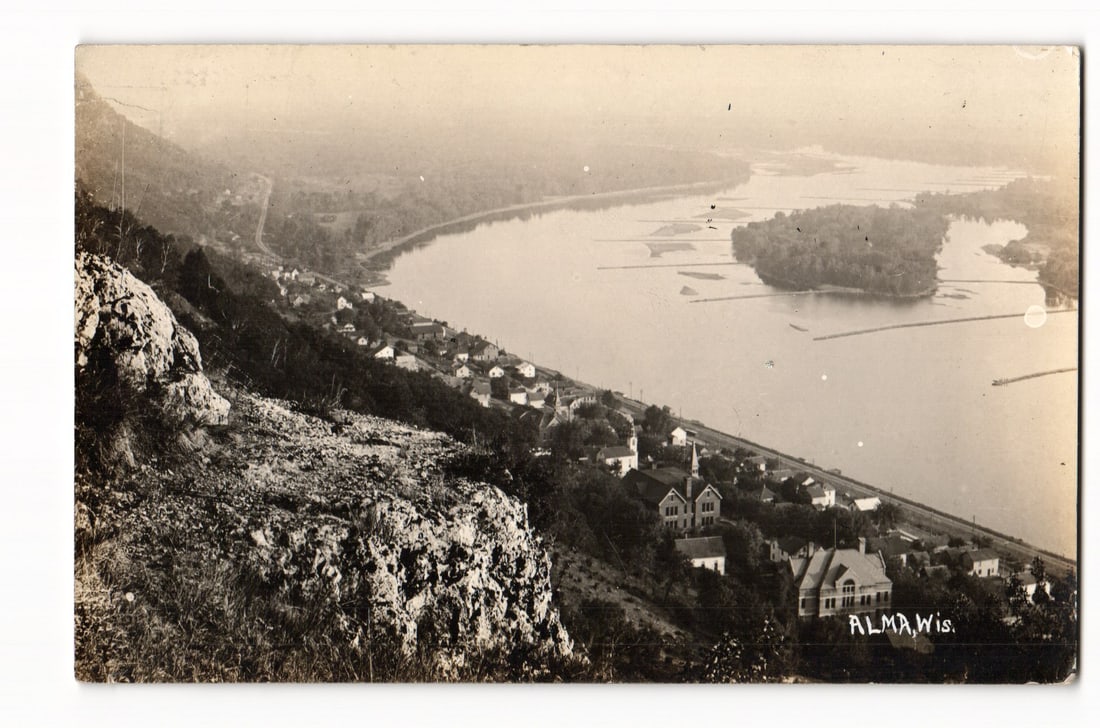 Alma, Wisconsin: Elevated Photographic View of Town, River, and Hills, Real Photo Postcard: A high-angle photographic view shows the town of Alma, Wisconsin, nestled along a wide river, with a prominent rocky and vegetated hillside in the immediate foreground. The town, characterized by its 