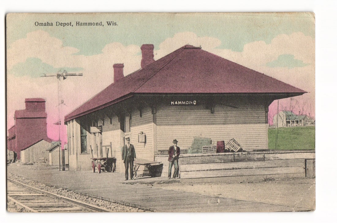 Colorized Postcard of the Omaha Depot in Hammond, Wisconsin, Featuring Two Men on the Platform: A colorized image depicting the Omaha Depot in Hammond, Wisconsin. The single-story depot building features a prominent hip roof with two chimneys and "HAMMOND" printed on its side. Two men in period