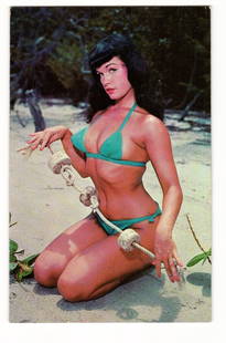 Postcard: "A Saucy Cutie Waits for You," Woman in Teal Bikini on Beach, Photo by Bunny Yeager: This printed color postcard features a woman with dark, styled hair and red lipstick, kneeling on a sandy beach. She wears a teal-colored two-piece bikini, consisting of a halter top and tie-side bott