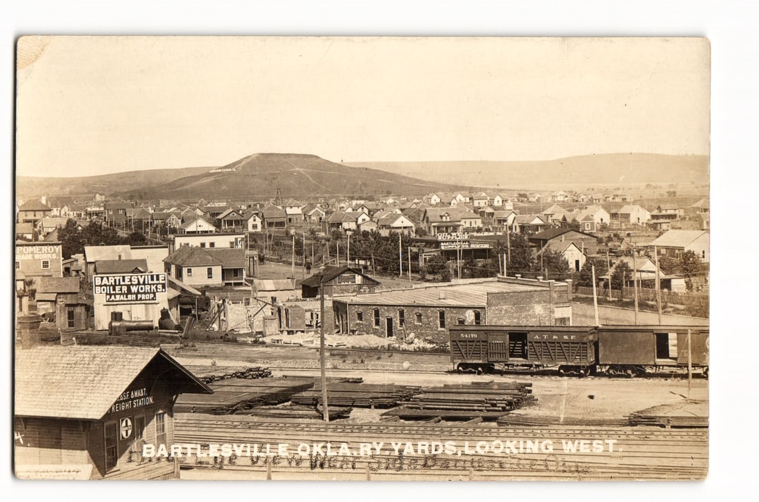 Bartlesville, Oklahoma, Railway Yards Looking West, Town View with Hill, Real Photo Postcard: A sepia-toned photograph showing an elevated, panoramic view of Bartlesville, Oklahoma, looking west. The foreground is dominated by railway yards, with multiple tracks, stacks of railroad ties or rai
