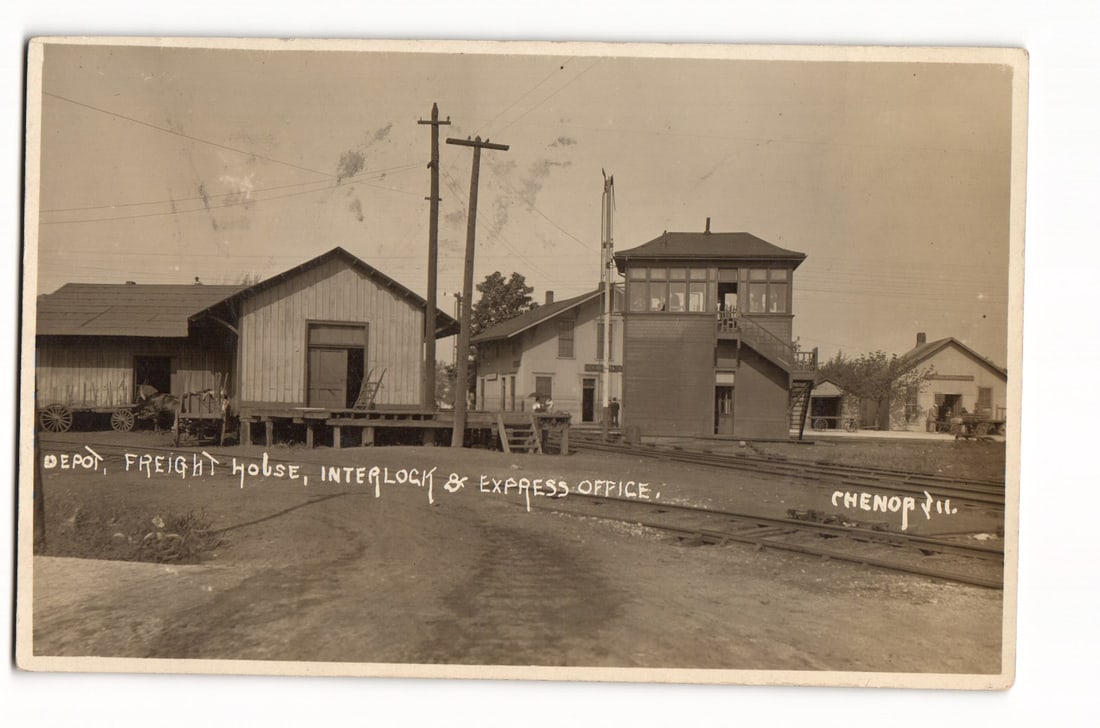 RPPC Chenoa, IL Railway Depot, Freight House, Interlock & Express Office, View from Tracks, 1910: A sepia-toned photograph depicts a collection of railway buildings in Chenoa, Illinois. From left to right, a freight house with a raised wooden platform and a horse-drawn wagon nearby, a smaller depo