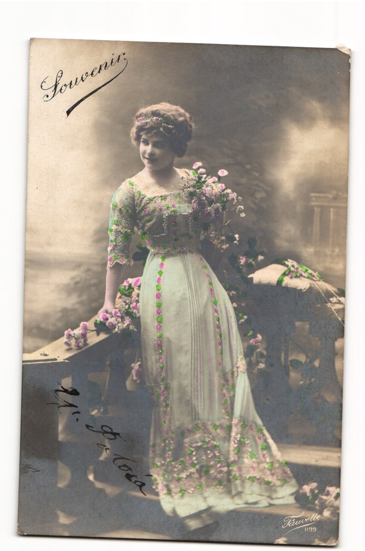 Souvenir Postcard: Elegant Woman with Flowers, Hand-Tinted Photographic Portrait, circa 1900s: A hand-tinted photographic postcard. The front features a full-length portrait of a woman in a light-colored, floor-length gown adorned with floral patterns highlighted in green and pink. She wears a