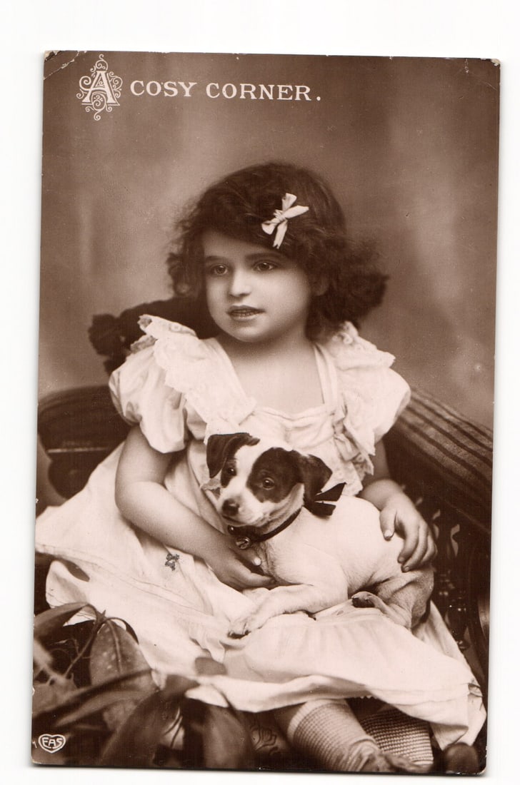Postcard: "A Cosy Corner," Young Girl with Bow in Hair Holding Small Dog, Sepia Photographic Print: A sepia-toned photographic print depicts a young girl with dark curly hair and a light-colored top bow, wearing a light-colored, ruffled dress. She is seated, holding a small, bi-colored (predominantl