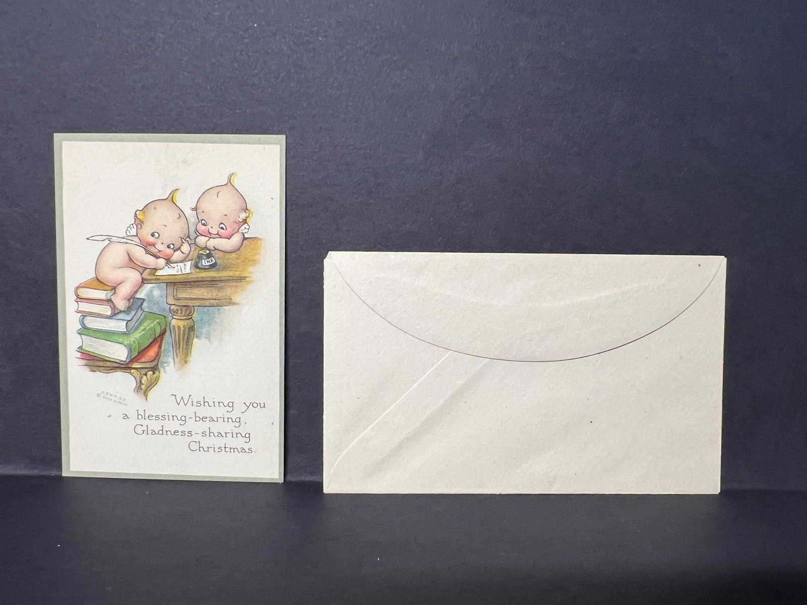 Antique Rose O'Neill Kewpie Christmas Greeting Card with Envelope – Blessing Message, c.1910s: Early 20th-century Christmas greeting card illustrated by Rose O’Neill featuring two Kewpie characters, one perched on a small stack of books and writing with a quill at a wooden desk, while the oth