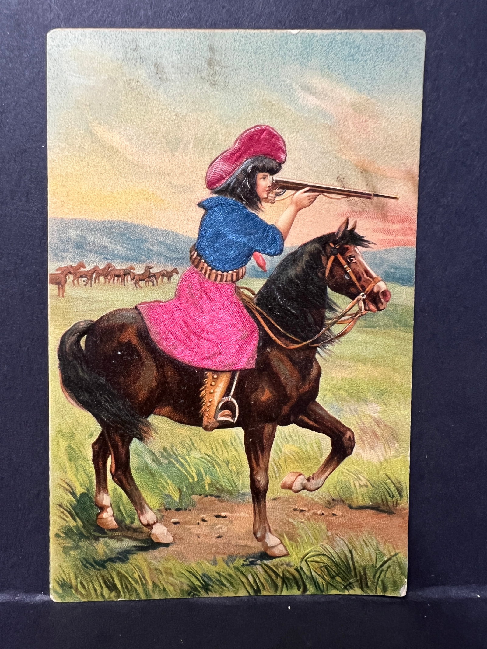 Embossed Novelty Postcard – Girl on Horseback with Applied Fabric Outfit, Made in Germany: Early 20th-century novelty postcard featuring a chromolithograph image of a woman on horseback aiming a rifle. The card includes applied textile embellishments simulating stitched fabric, affixed to t