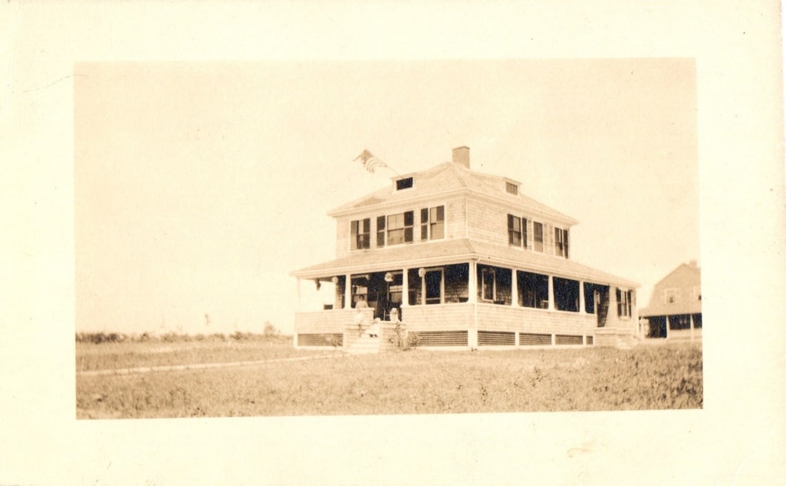 Sepia Real Photo Postcard Depicting a Large Shingled House with Wrap-Around Porch and Flying Flag: A large, two-story wooden house with shingled siding and a wide wrap-around porch. A flag flies from a pole on the roof of the house. The house features numerous windows on both levels and multiple co