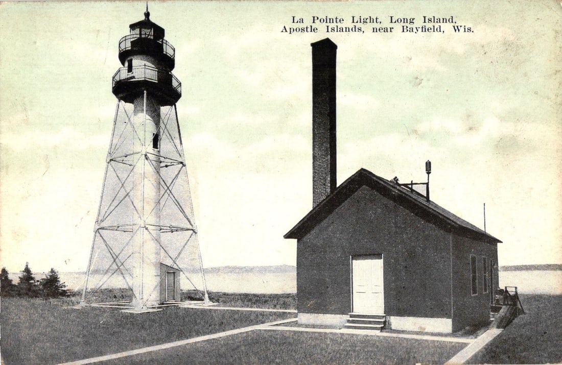 La Pointe Light, Apostle Islands, Bayfield, Wisconsin postcard from 1922, featuring lighthouse.: A tall, white, open-framework lighthouse stands prominently on the left, adjacent to a dark, single-story building featuring a tall brick chimney on the right. The lighthouse is a pyramidal skeletal s