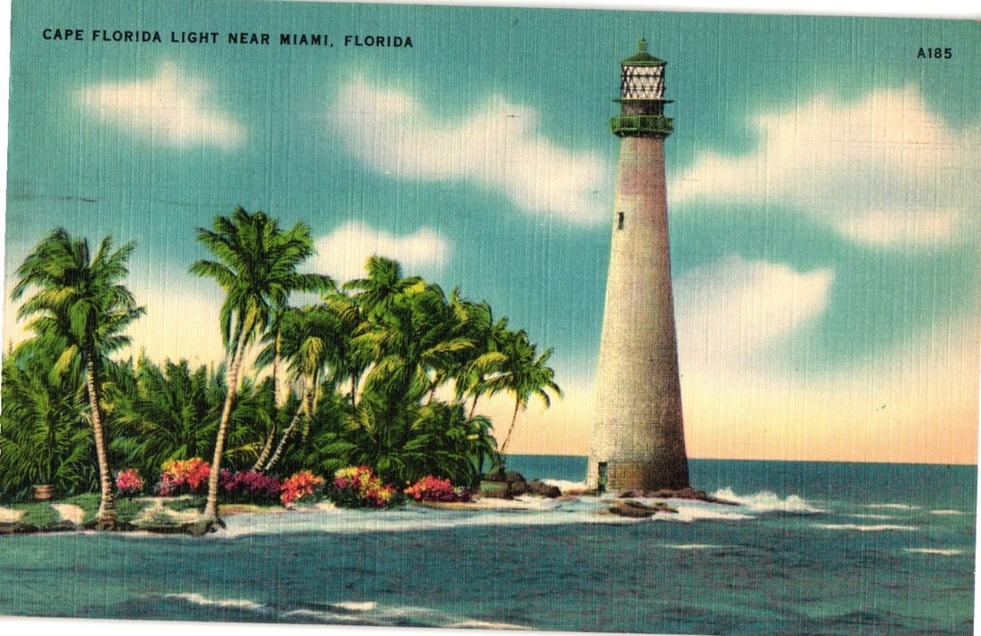 Cape Florida Light Near Miami, Florida: 1949 Lighthouse and Palm Trees Coastal View postcard.: A tall, tapering lighthouse stands on a rocky shore at the edge of calm ocean waters. To the left, a cluster of palm trees and flowering shrubs occupies a small island. White clouds are scattered acro