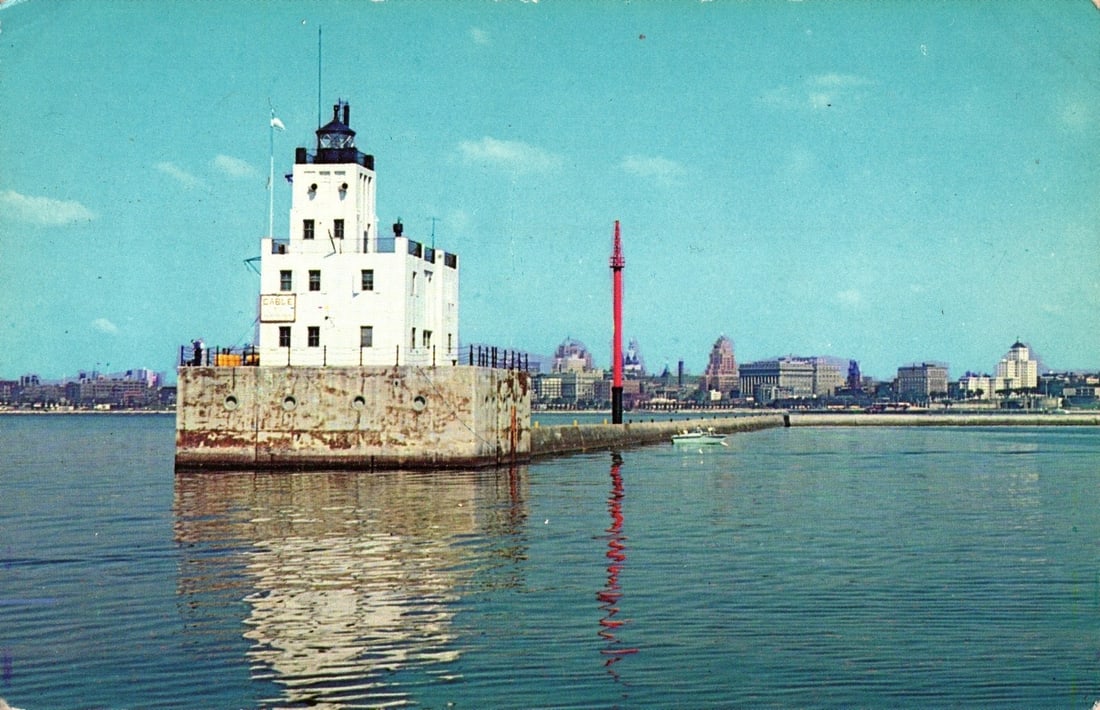 Milwaukee Breakwater Light, Port of Milwaukee, Wisconsin postcard, City Skyline, 1960s: A prominent white multi-story lighthouse structure with a dark cap and flag pole stands on a robust concrete base in a body of water. To its right, a tall red pole with a small platform is visible. A 