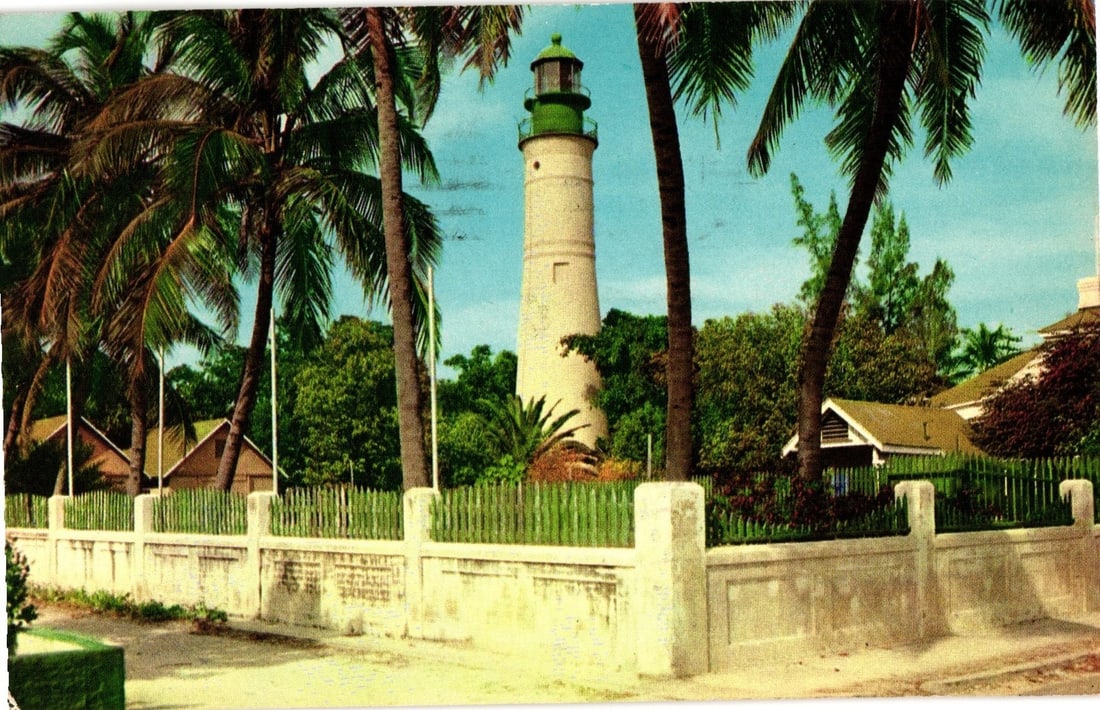 Scenic View of Famous Lighthouse, Key West, Florida, framed by palm trees, 1957 Postmark postcard: A tall, white cylindrical lighthouse with a green lantern room dominates the central composition, framed by several prominent palm trees with large fronds. Lush green vegetation and parts of small bui