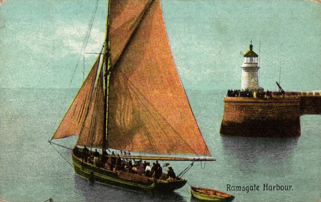 Early 20th century postcard depicting a sailboat near a lighthouse in Ramsgate Harbour, England.: A large sailboat with a reddish-brown gaff sail and a dark hull carries many figures on deck, positioned prominently in the left foreground. A smaller, light-colored dinghy floats behind it. To the ri