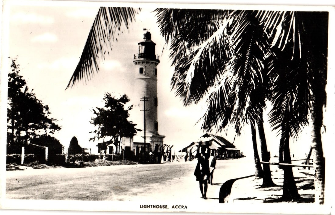Vintage Real Photo postcard depicting the Lighthouse in Accra, Ghana, with palm trees and figures.: The Lighthouse in Accra, Ghana, is prominently featured in this black and white real photo postcard. The tall, cylindrical lighthouse stands on the left, accompanied by an adjacent structure at its ba