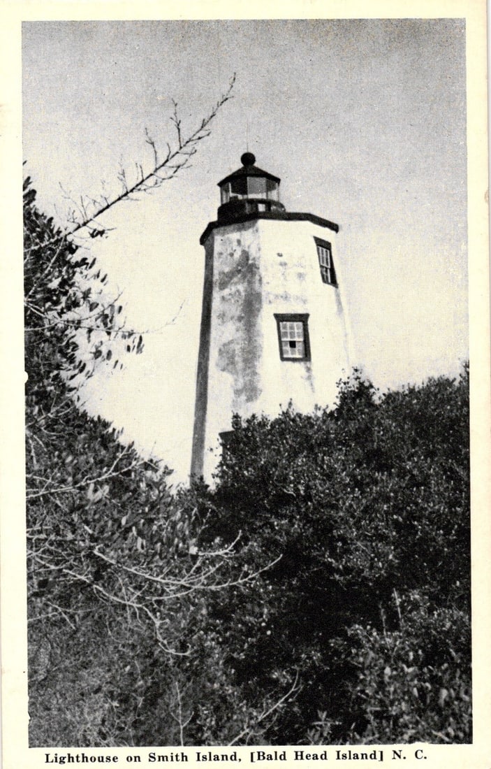 Black and white photographic postcard of the Lighthouse on Smith Island, Bald Head Island, N. C.: A lighthouse stands prominently amidst dense, low-lying vegetation. The perspective is from below, looking up at the structure, with branches of a tree extending into the upper left quadrant. The ligh