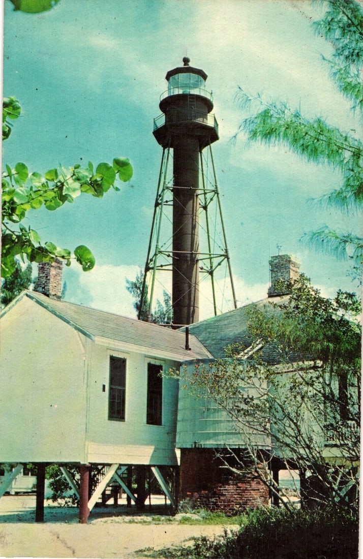 Historic Sanibel Island Lighthouse and Keeper's Quarters postcard, Florida, mid-20th century: A tall, skeletal lighthouse tower, dark brown or black, rises into a light blue sky with scattered clouds. The lighthouse features a prominent lantern room at its apex. Below and to the left of the to
