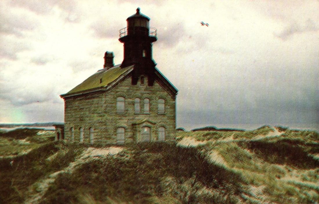 Scenic postcard of the historic North Light, Block Island, Rhode Island, granite-built in 1867: The North Light, a historic granite lighthouse, stands atop a sandy dune. A single seagull is visible flying in the overcast sky. The lighthouse is positioned slightly left of center, with wind-swept 