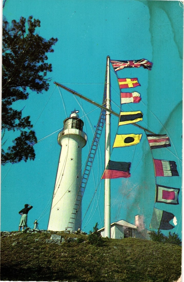 Bermuda's Gibbs Hill Lighthouse with Signal Flags and a Figure on Hilltop, Color Postcard: Gibbs Hill Lighthouse, a white cylindrical tower with a lantern room, stands on a grassy hilltop. A tall flagpole to its right displays multiple signal flags against a clear blue sky. A small building