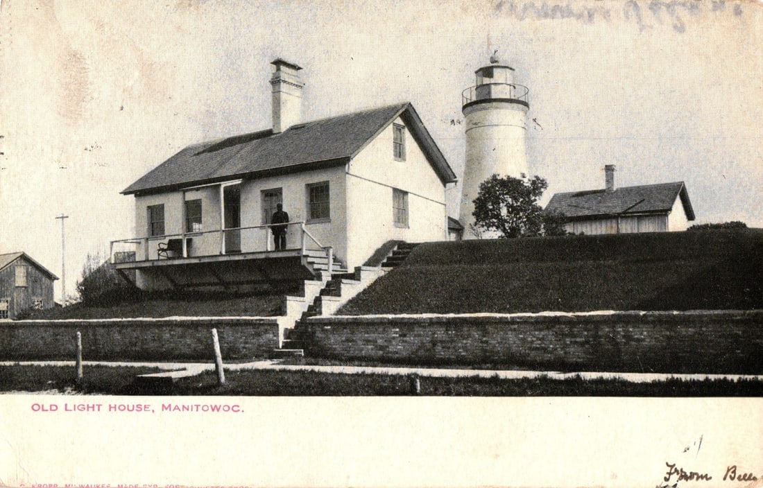 Manitowoc, Wisconsin: Historic Lighthouse and Keeper's House, 1905-era photographic postcard: A historic lighthouse, cylindrical and white, stands next to a two-story white house with a gabled roof and chimney. A figure is visible on the house's porch, which features a railing and steps leadin
