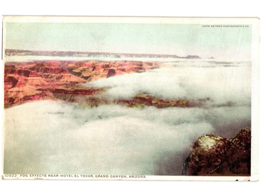 Grand Canyon, Arizona, Fog Effects Near Hotel El Tovar, 1912 Photochrome Postcard: A sweeping view of the Grand Canyon, Arizona, largely enveloped in thick white fog and clouds. Red-hued canyon walls are visible above and through the fog in the background and midground. A jagged, ro